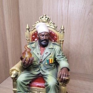 May include: A detailed figurine of a bearded individual wearing a light-coloured turban and a green military-style jacket, seated on an ornate gold throne with red upholstery. The figure holds a translucent orange flame and wears a gold ring.