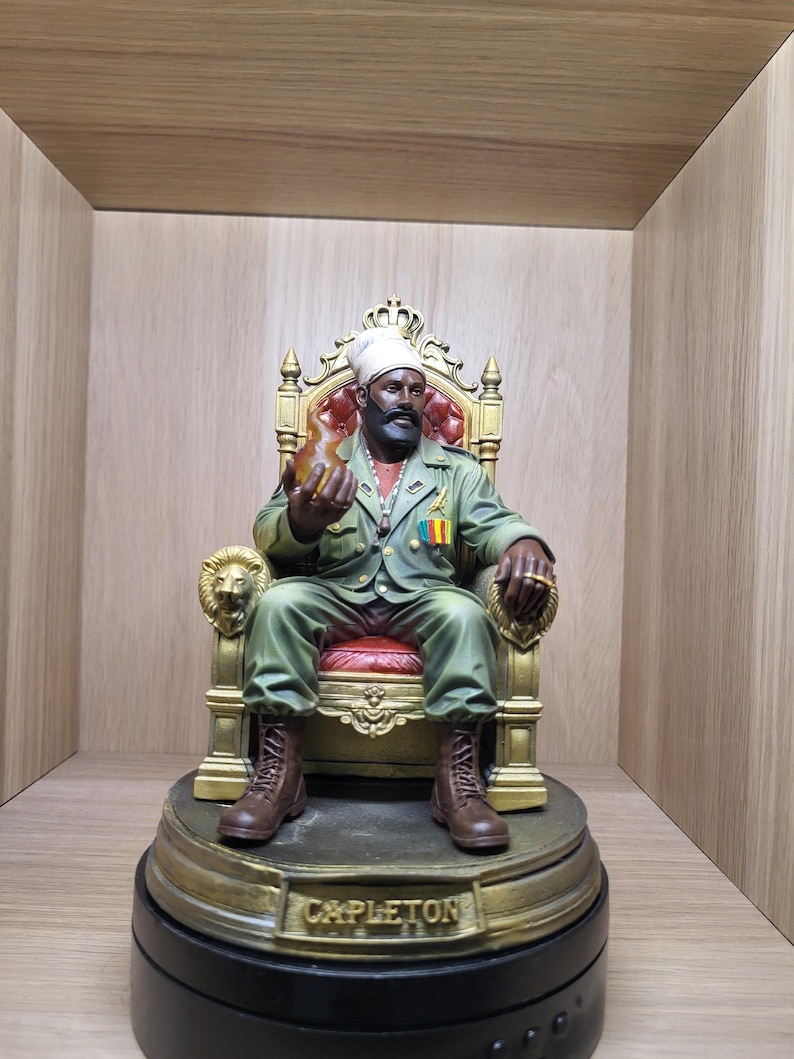 May include: A detailed figurine of a bearded person wearing a white head wrap and olive green military-style jacket, seated on an ornate gold throne with red upholstery. The figure holds a translucent orange flame and has the name CAPLETON on the base.
