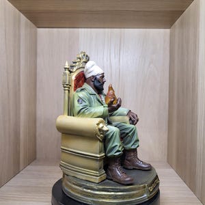 May include: A figurine of a bearded person wearing a white turban, olive green jacket, and brown boots, seated on a gold-coloured throne with lion-head armrests. The figure holds a translucent orange flame-shaped object in one hand.