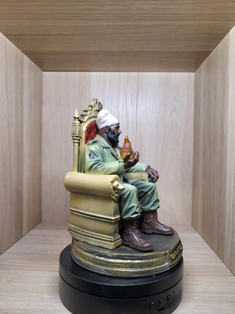 May include: A figurine of a bearded person wearing a white turban, olive green jacket, and brown boots, seated on a gold-coloured throne with lion-head armrests. The figure holds a translucent orange flame-shaped object in one hand.