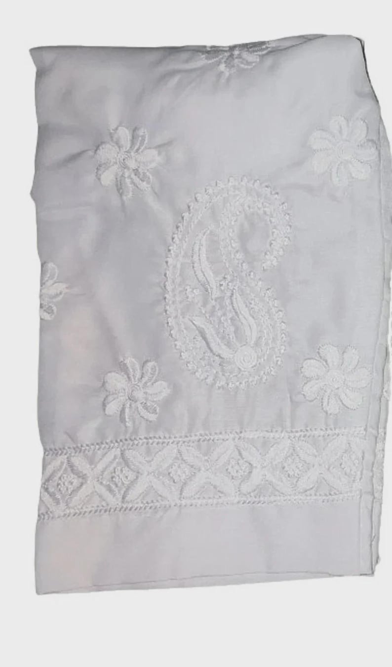 Puede incluir: Tejido blanco con intrincados bordados florales y de cachemira. El dise&ntilde;o presenta costuras detalladas, incluyendo flores y un motivo central de cachemira. La tela est&aacute; doblada, mostrando el delicado trabajo de bordado.