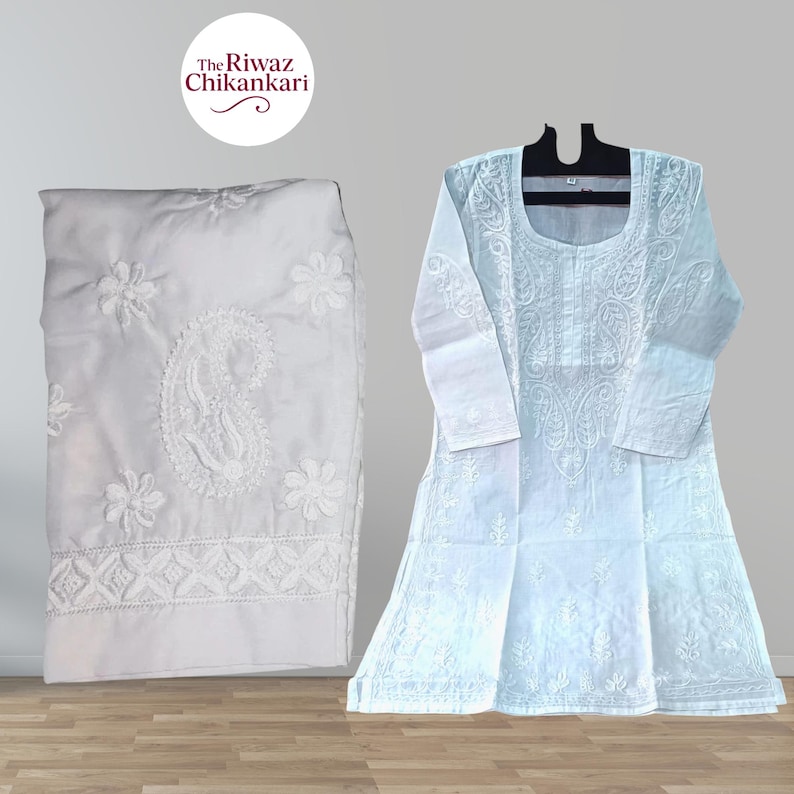Puede incluir: Conjunto de kurta y pantal&oacute;n blancos de Chikankari. La kurta presenta intrincados bordados florales y de cachemira. El pantal&oacute;n est&aacute; doblado y tambi&eacute;n muestra bordados detallados. El conjunto se exhibe sobre una superficie de madera.