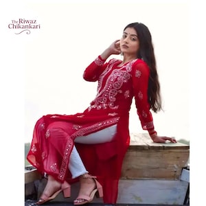 May include: A long, red tunic with intricate white embroidery and embellishments. The tunic features long sleeves and a high slit, paired with white pants. The text "The Riwaz Chikankari" is in the upper left corner. The outfit is worn with pink heels.