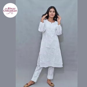 May include: White Chikankari kurta and pants set. The long-sleeved kurta features intricate floral embroidery around the neckline and down the front. The matching pants also have embroidered details. The brand logo "The Riwaz Chikankari" is in the upper left corner.
