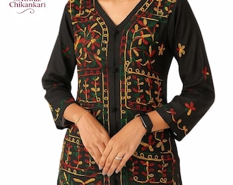 Puede incluir: Túnica negra de manga larga con bordados florales coloridos. La túnica presenta un cuello en V y cierre de botones. El bordado incluye patrones florales rojos, amarillos y verdes. La marca "The Riwaz Chikankari" es visible en la esquina superior izquierda.