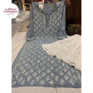以下が含まれることがあります： 精巧な刺繍とミラーワークが施された、水色のチカンカリのスーツセット。セットには、長いチュニックとカットワークのディテールが施された白いパンツが含まれています。「The Riwaz Chikankari」のブランド名が表示されています。