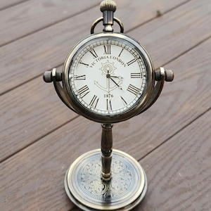 May include: Antique brass-colored desk clock with a round face, featuring Roman numerals and the words "VICTORIA LONDON" and "1876". The clock has a nautical anchor and wheel design. It stands on a decorative base.