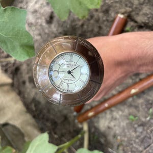 May include: A vintage-style walking cane with a clock face. The clock has Roman numerals and a cream-colored face. The cane's handle is a bronze color, and the shaft is brown. The cane is resting on a natural surface.