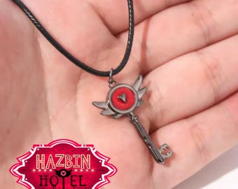 May include: A black cord necklace with a gunmetal key pendant. The key has a red heart-shaped centre, silver wings, and a keyhole-shaped end. The image also includes a red and white logo that says "HAZBIN HOTEL".