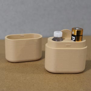 May include: Two beige wooden containers. One is an oval-shaped box. The other is a cylindrical container with a small bottle and a gold and black battery inside. The containers are on a neutral background.
