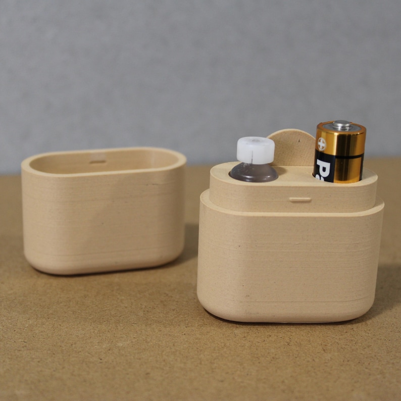 May include: Two beige wooden containers. One is an oval-shaped box. The other is a cylindrical container with a small bottle and a gold and black battery inside. The containers are on a neutral background.