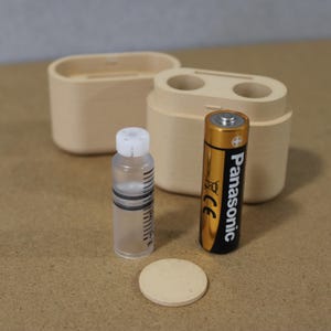 May include: A wooden container with two circular openings, a small clear bottle with a white cap, a beige disc, and a gold and black Panasonic AA battery. The items are arranged on a light brown surface.