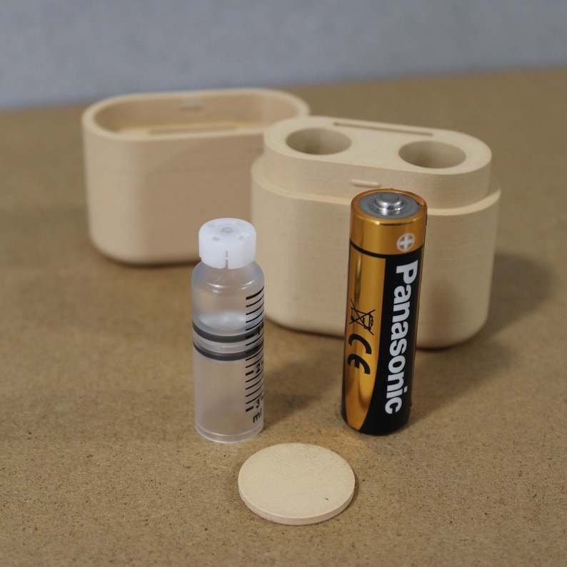 May include: A wooden container with two circular openings, a small clear bottle with a white cap, a beige disc, and a gold and black Panasonic AA battery. The items are arranged on a light brown surface.