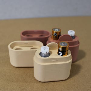 May include: Four small, light-colored battery holders in varying shapes. Two holders contain AA batteries. The holders are in shades of beige and terracotta. The background is a neutral, light brown.