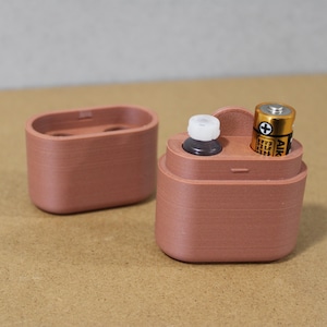 May include: A terracotta-colored, 3D-printed container with a separate lid. The container holds a small bottle with a white cap and a AA battery. The lid has two small holes. The items are on a neutral background.