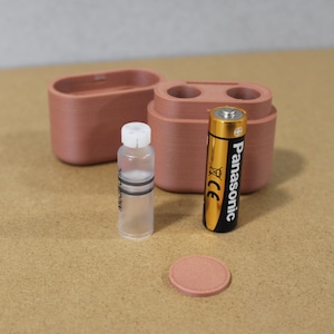 May include: A collection of small, terracotta-colored containers and accessories. Includes a small bottle with a white cap, a Panasonic AA battery, and a round terracotta-colored disc. The items are arranged on a light brown surface.