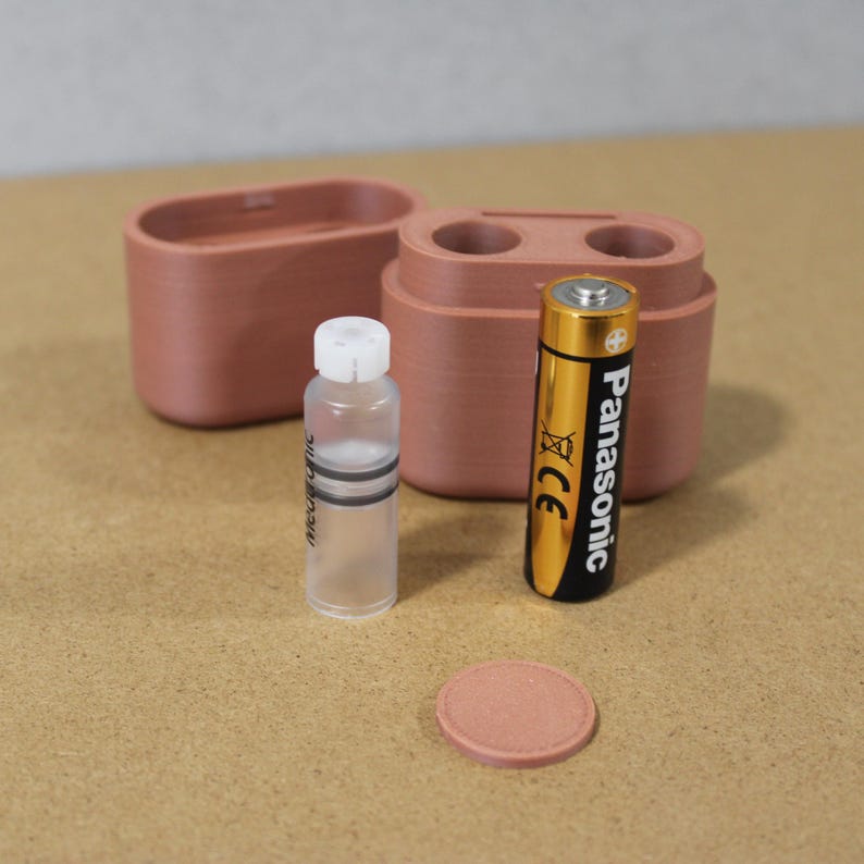May include: A collection of small, terracotta-colored containers and accessories. Includes a small bottle with a white cap, a Panasonic AA battery, and a round terracotta-colored disc. The items are arranged on a light brown surface.