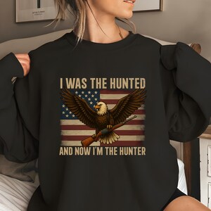 Pode incluir: Camisola preta com uma águia segurando um rifle, contra uma bandeira americana. O texto diz: "I WAS THE HUNTED AND NOW I'M THE HUNTER."