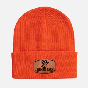 May include: Bright orange beanie with a folded brim. A rectangular patch with the text "FLEMING FARM" and a goat graphic is sewn on the front. The beanie is made of a knitted material.