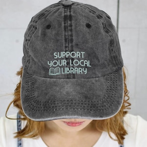 May include: A distressed charcoal gray baseball cap with the embroidered text "SUPPORT YOUR LOCAL LIBRARY" in light green. The cap features a curved brim and a classic six-panel design, perfect for casual wear.