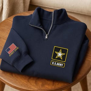 May include: A navy blue quarter-zip sweatshirt featuring an embroidered U.S. Army logo with a yellow-bordered black square and a white star on the chest. The sleeve displays an embroidered American flag above the text JOHN DOE in yellow thread.