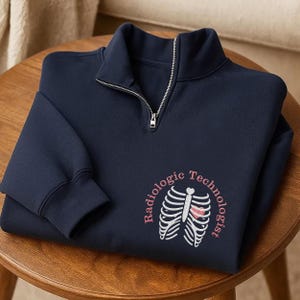 May include: Navy blue sweatshirt with a quarter-zip collar. The sweatshirt features the words "Radiologic Technologist" in pink, encircling a white rib cage graphic with a pink heart.