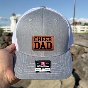 May include: A grey and white trucker hat with the words "CHEER DAD" on a brown leather patch. The hat has a Richardson 112 tag.
