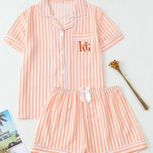 Puede incluir: Conjunto de pijama a rayas melocotón y blanco. La parte superior de manga corta tiene una botonadura, un bolsillo en el pecho con iniciales bordadas y ribetes azul claro. Los pantalones cortos a juego tienen un lazo blanco. Un pequeño ramo de flores secas está al lado del pijama.