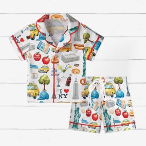 Puede incluir: Conjunto de pijama blanco con tem&aacute;tica de la ciudad de Nueva York. El conjunto incluye una camisa de manga corta con botones y pantalones cortos a juego. El estampado presenta taxis amarillos, manzanas rojas, la Estatua de la Libertad y el texto "I ❤️ NY."