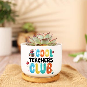 May include: A white ceramic planter with the text "COOL TEACHERS CLUB" in colorful letters. The planter holds a succulent plant with green and red leaves. The planter sits on a wooden coaster and a burlap mat.