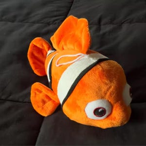 May include: A plush toy clownfish in bright orange with white and black stripes. The stuffed animal has large, round eyes with red and black details, and orange fins. The toy is designed to resemble a clownfish.