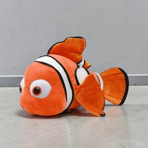 May include: A plush toy fish in orange, white, and black, resembling a clownfish. The toy has large, round eyes and fins. The fish is lying on a gray surface.