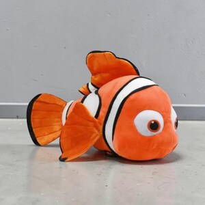 May include: A plush orange clownfish toy with white stripes and large, expressive eyes. The stuffed animal has orange fins and a black outline, resting on a gray surface. A children's toy.