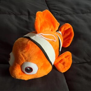 May include: A plush toy fish in bright orange with white and black stripes. The fish has large, round eyes and orange fins. The toy is made of soft material and is designed to resemble a clownfish.