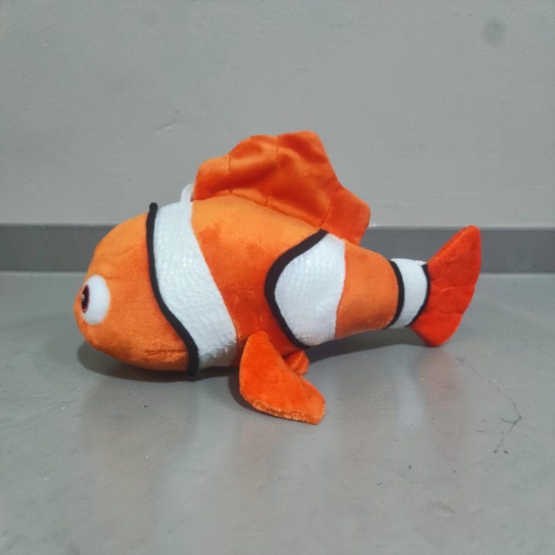 May include: A plush toy clownfish in orange, white, and black. The stuffed animal has a large dorsal fin, a rounded body, and a tail fin. The toy is designed to resemble a clownfish.