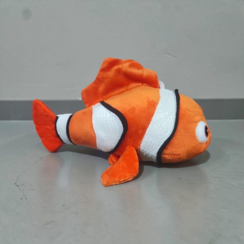 May include: A plush toy clownfish in orange, white, and black. The stuffed animal has a large orange fin on top and a small orange fin on the bottom. The toy has a white stripe outlined in black around its body and a red eye.