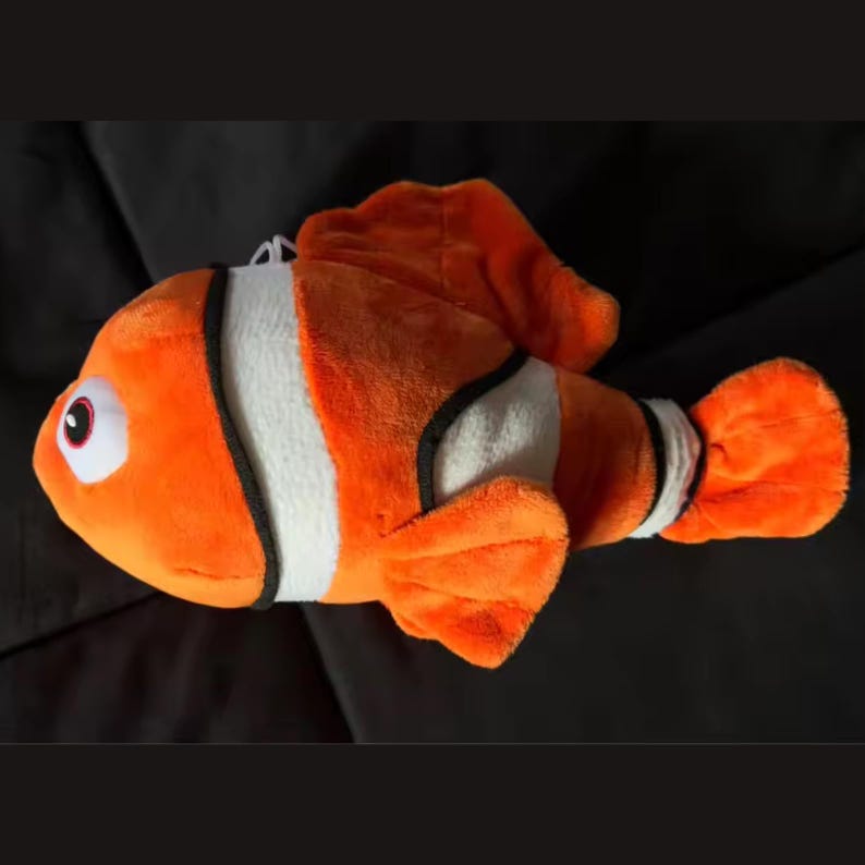 May include: Orange plush toy fish with white and black stripes. The toy has large, round eyes and fins. The fish is lying on a black surface.