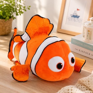 May include: A plush orange and white clownfish toy with large eyes and black outlines. The stuffed animal has a black tail and fins, and is lying on a light-colored surface. The toy is a children's item.