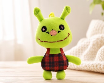 May include: A plush green creature with stitched features, including black cross-shaped eyes and a smiling mouth. It wears red and black plaid overalls with red straps. The toy has two antennae and a small yellow tuft of hair.