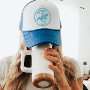 May include: A white and blue trucker hat with the text "GOODBYE EARL" and a turquoise graphic of a person riding a horse. The hat is worn by a person holding a white mug with a wooden base.