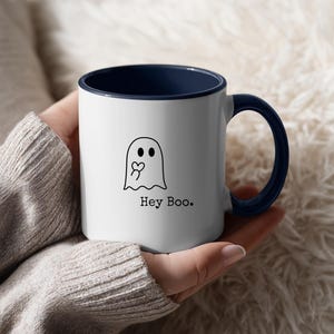 May include: White ceramic mug with a navy blue interior and handle. The mug features a black line drawing of a ghost holding a heart, with the text "Hey Boo." in a simple font. The mug is held in a person's hands.