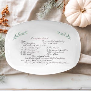 May include: White oval serving platter with a handwritten recipe for pumpkin bread. The platter is decorated with green leaf sprigs and sits on a neutral-toned linen cloth. A white pumpkin and pine cones are in the background.