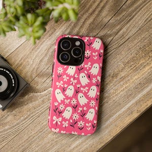 May include: A pink smartphone case decorated with a pattern of white ghosts, flowers, and bows. The case rests on a wooden surface, with a camera and plant in the background. The case is designed for a smartphone.
