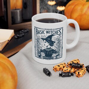 Könnte beinhalten: Weiße Keramiktasse mit einer schwarz-blauen Grafik einer Frau mit Hexenhut und dem Text "BASIC WITCHES BOOKCLUB". Die Tasse ist mit einer dunklen Flüssigkeit gefüllt. Halloween-Süßigkeiten sind um die Tasse herum verstreut.