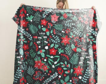 Könnte beinhalten: Eine schwarze Decke mit einem festlichen Blumenmuster. Das Design zeigt rote Weihnachtssternblumen, grüne Blätter, weiße Beeren und andere botanische Elemente. Die Decke wird hochgehalten, um das volle Muster zu zeigen.