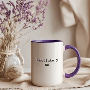 Puede incluir: Taza de cerámica blanca con interior y asa morados. La taza tiene el texto "Immediately No." en negro. Un jarrón de flores moradas secas está detrás de la taza sobre una superficie neutra.