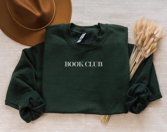 May include: A dark green sweatshirt with the words "BOOK CLUB" printed in white. The sleeves are folded. A brown hat and dried flowers are in the background. The sweatshirt is made of a soft material.