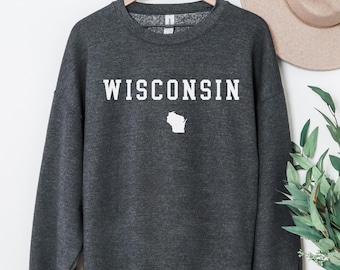 May include: A dark gray sweatshirt with the word "WISCONSIN" in white block letters and a white outline of the state. The sweatshirt is hanging on a wooden hanger against a white background.