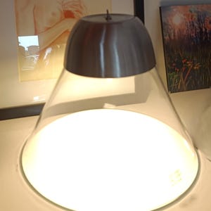 Peut inclure: Une lampe de table moderne avec un abat-jour en forme de dôme en métal brossé et un diffuseur en verre conique transparent. La lampe émet une lumière vive et chaude. La lampe est sur une surface blanche.