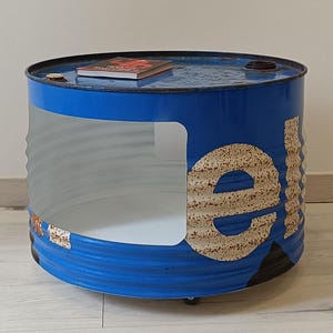 Puede incluir: Una mesa de centro única hecha de un barril de metal azul reciclado. El barril tiene una sección recortada, que revela un interior blanco. El exterior tiene un aspecto envejecido y rústico con la palabra "fuel" visible. Un libro descansa sobre la mesa.